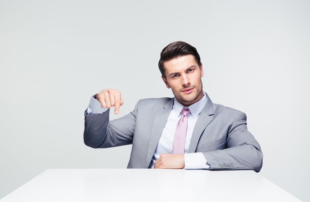 Confident businessman sitting at the table pointing finger down over gray background. Loooking at camera